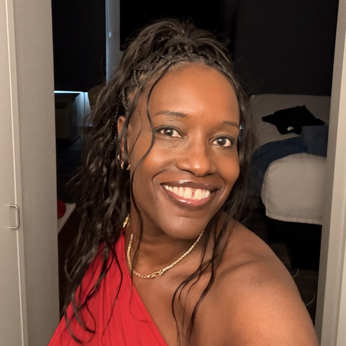 Close-up portrait of a smiling Black woman with braided hair and a red top, wearing a gold necklace, taking a selfie in a doorway.
